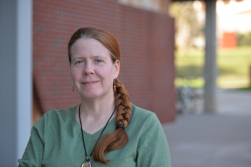 Photo of Dr. Barbara Stock. Philosophy Program Coordinator. Dr. Stock has long red hair which is pulled back; she is wearing a short sleeved green t-shirt. and is standing outside of the SAC against a red brick wall.
