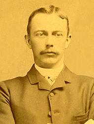 Olof Hanson in his school picture from 1886. The image appears to have an orange tone. Hanson is wearing a suit and is sporting a mustache.