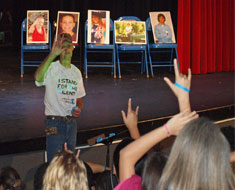 Image: Kirk Smalley signs "I Love You" to the audience after giving a talk on October 27 at Theatre Malz on the effects of bullying. Photographs of children who have committed suicide as a result of bullying are displayed on the stage behind him. (Photo: Susan Flanigan)