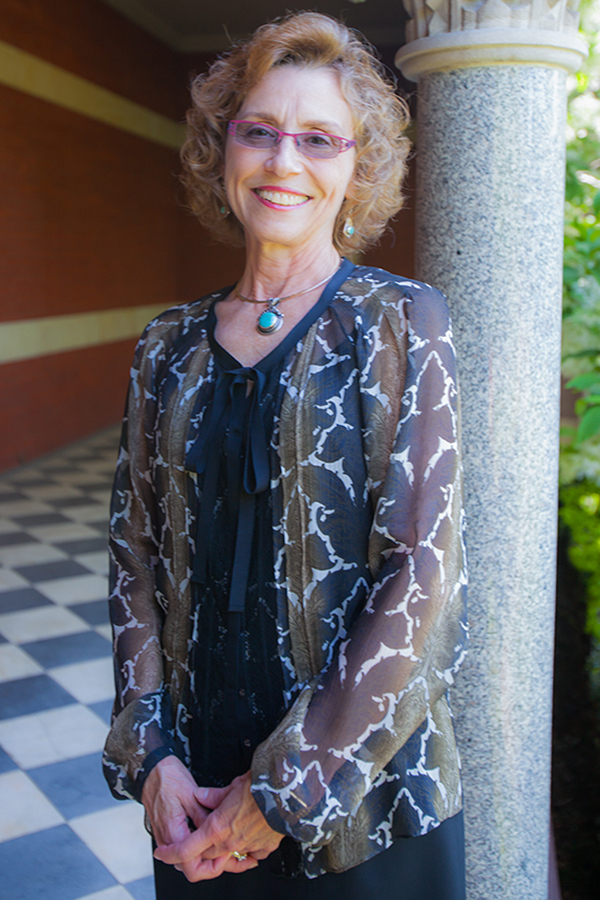 Dr Carol Erting standing outside by a column, gauzy gray top, pink glasses, chin length curly brown hair