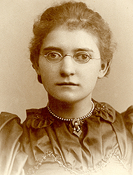 Agatha Tiegal Hanson in her class photo from 1893. The image has a grayish tone. She's wearing a dark dress and a necklace. Her glasses have small roundish frames, and her hair is pulled back
