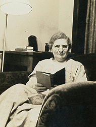 Agatha Tiegal Hanson, sitting in a comfortable chair, with a book in her hand. She is looking at the camera. A lamp is in the background.