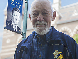 Jack Gannon standing near the light pole banner that bears his 1959 class photo. He's wearing his '59 class jacket and is smiling, with a bearded face.