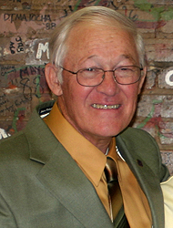 Jack Gannon, clean-shaven and smiling against the brick wall in the Peikoff Alumni House. He's wearing an olive-green suit jacket, goldish colored shirt and a tie with goldish hues and brown.