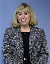 Claire Bugen, wearing black pants and blouse, with a black and white patterned jacket. She's sitting in front of a blue background