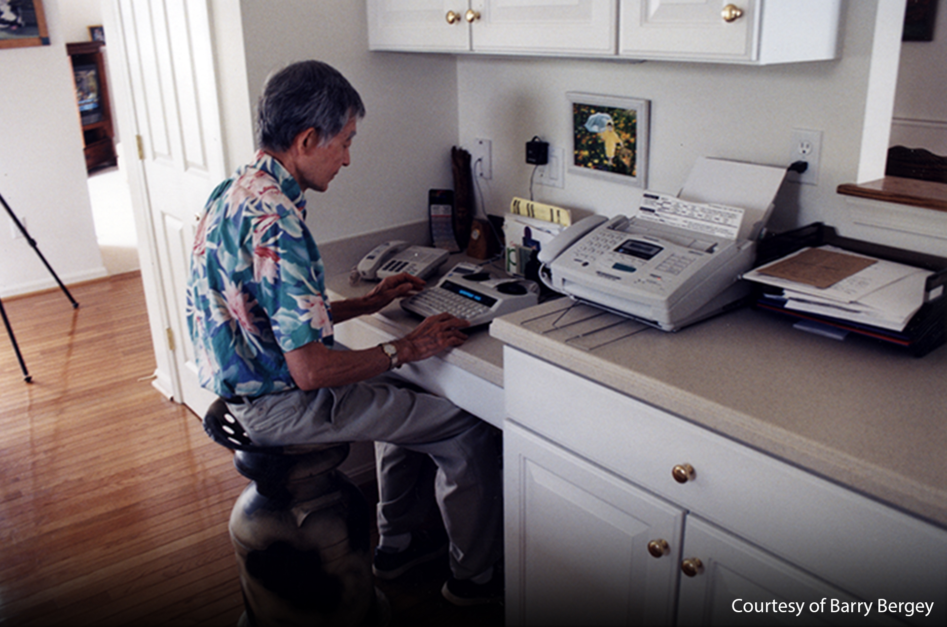 A man is sitting what appears to be a desk corner in a kitchen - he's using a TTY. There are some papers and a fax machine on the desk as well.