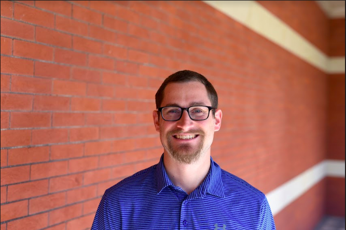A picture of a smiling caucasian man with brown hair and black eyeglasses in a blue jacket standing in the front of a red brick wall.