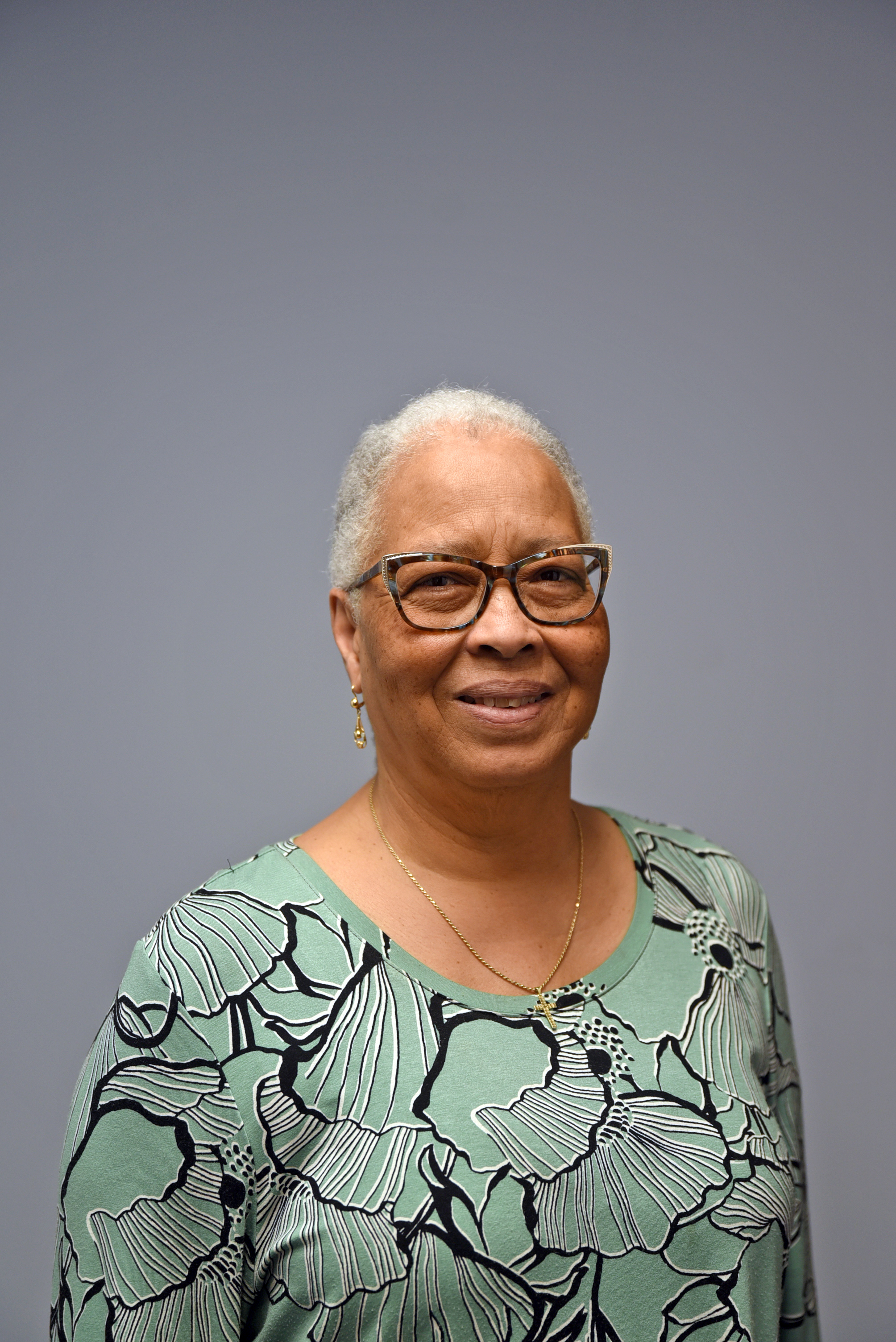 A picture of a smiling brown-skinned woman with white hair and brown eyeglasses in a flower-designed green jacket standing in the front of a grey wall.