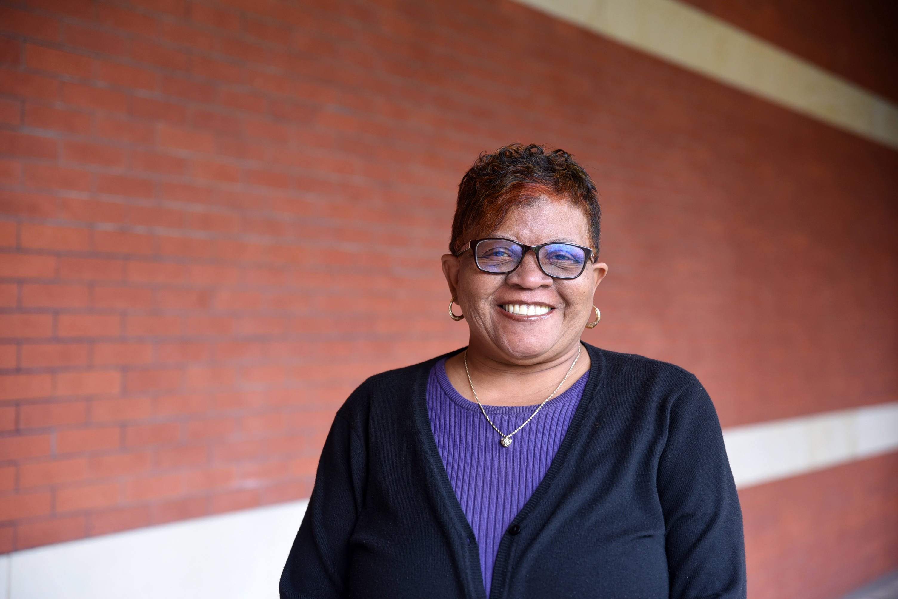 A picture of a smiling dark-skinned woman with black-brown mixed hair and black eyeglasses standing in the front of a red brick wall. She wears a purple jacket along with a dark blue thin coat.