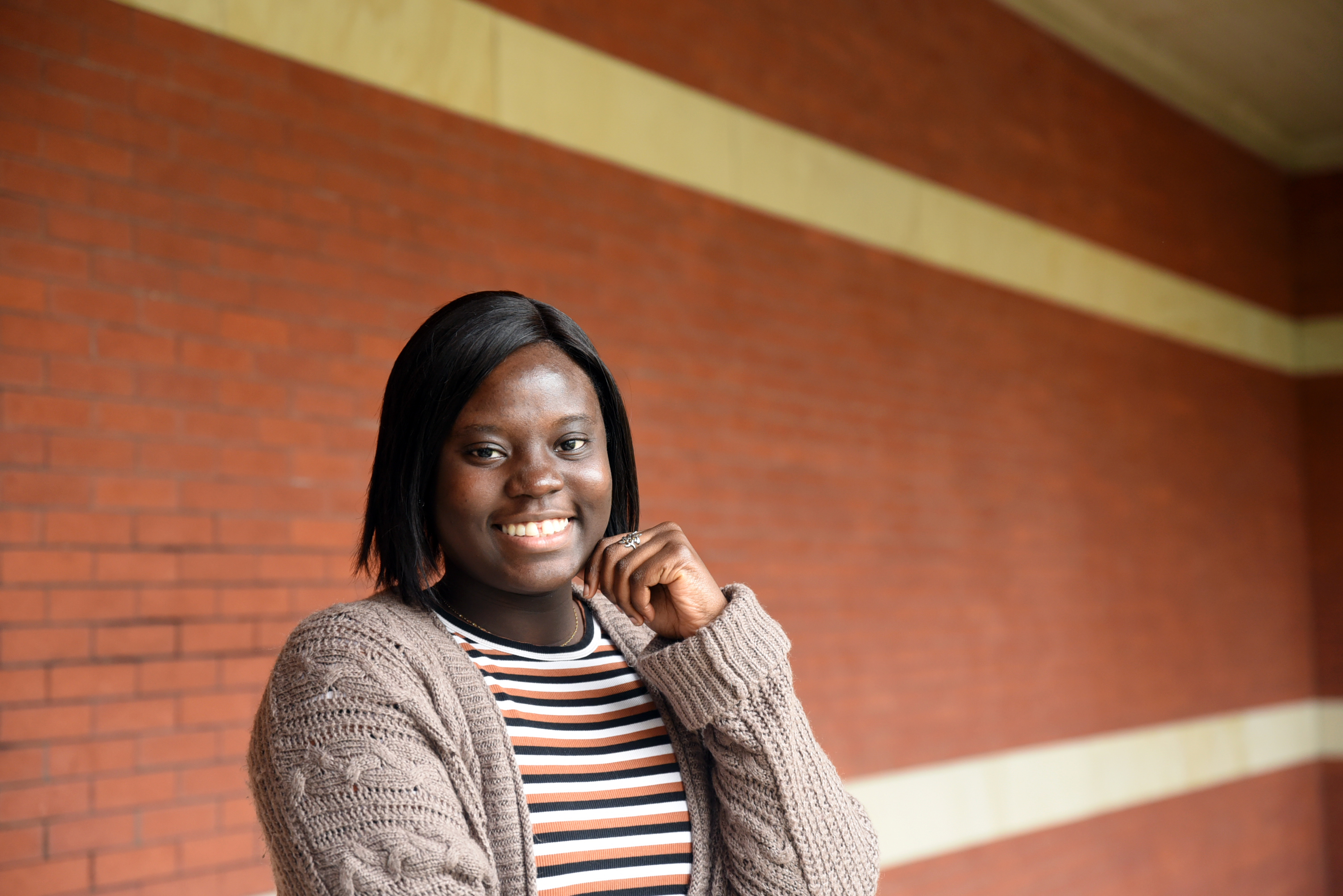 A picture of a smiling dark-skinned woman with black hair in a striped shirt along with a light-brown sweater standing in the front of a red brick wall.