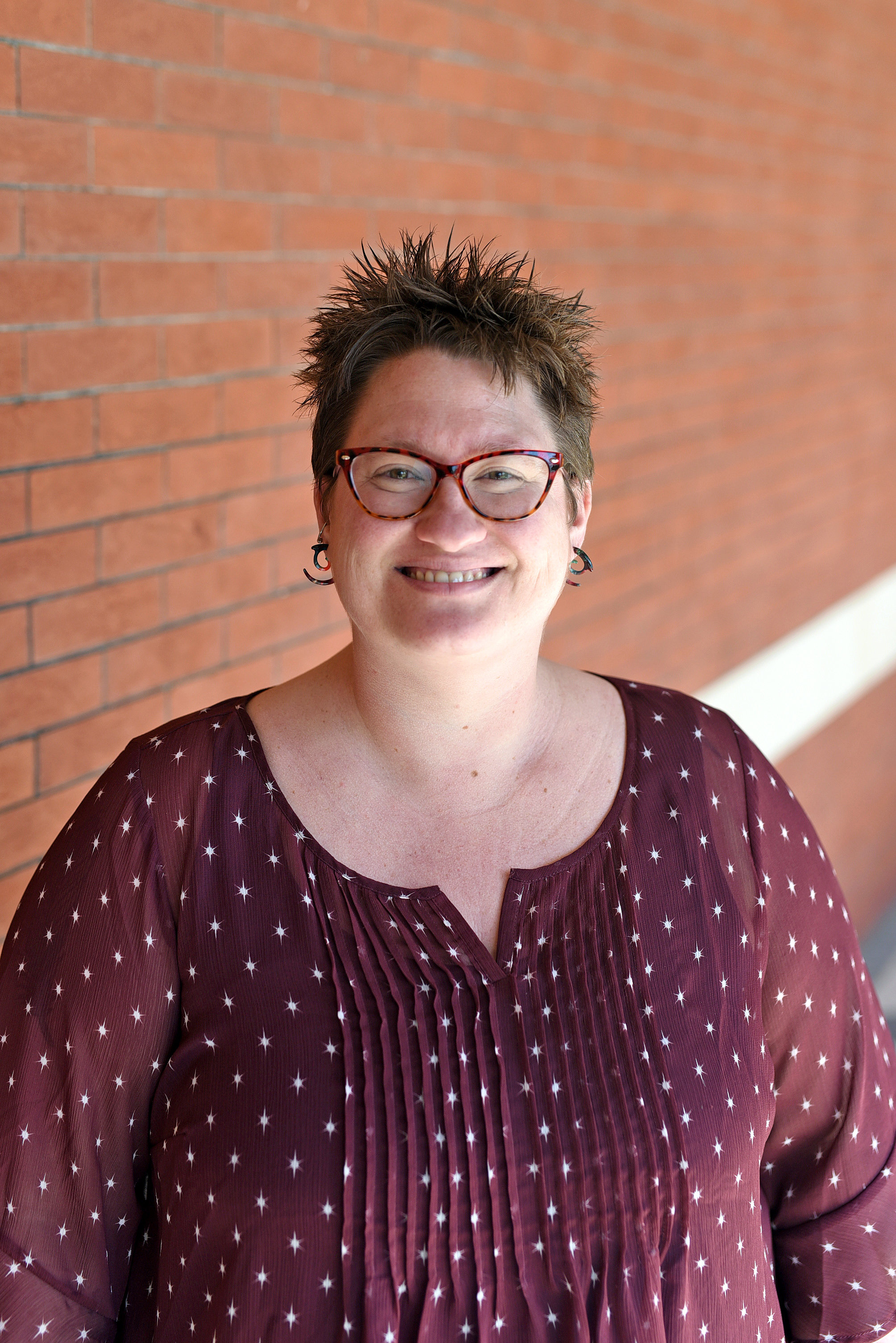 A picture of a smiling caucasian woman with short brown hair and red eyeglasses in a wine-red spotty jacket standing in the front of a red brick wall