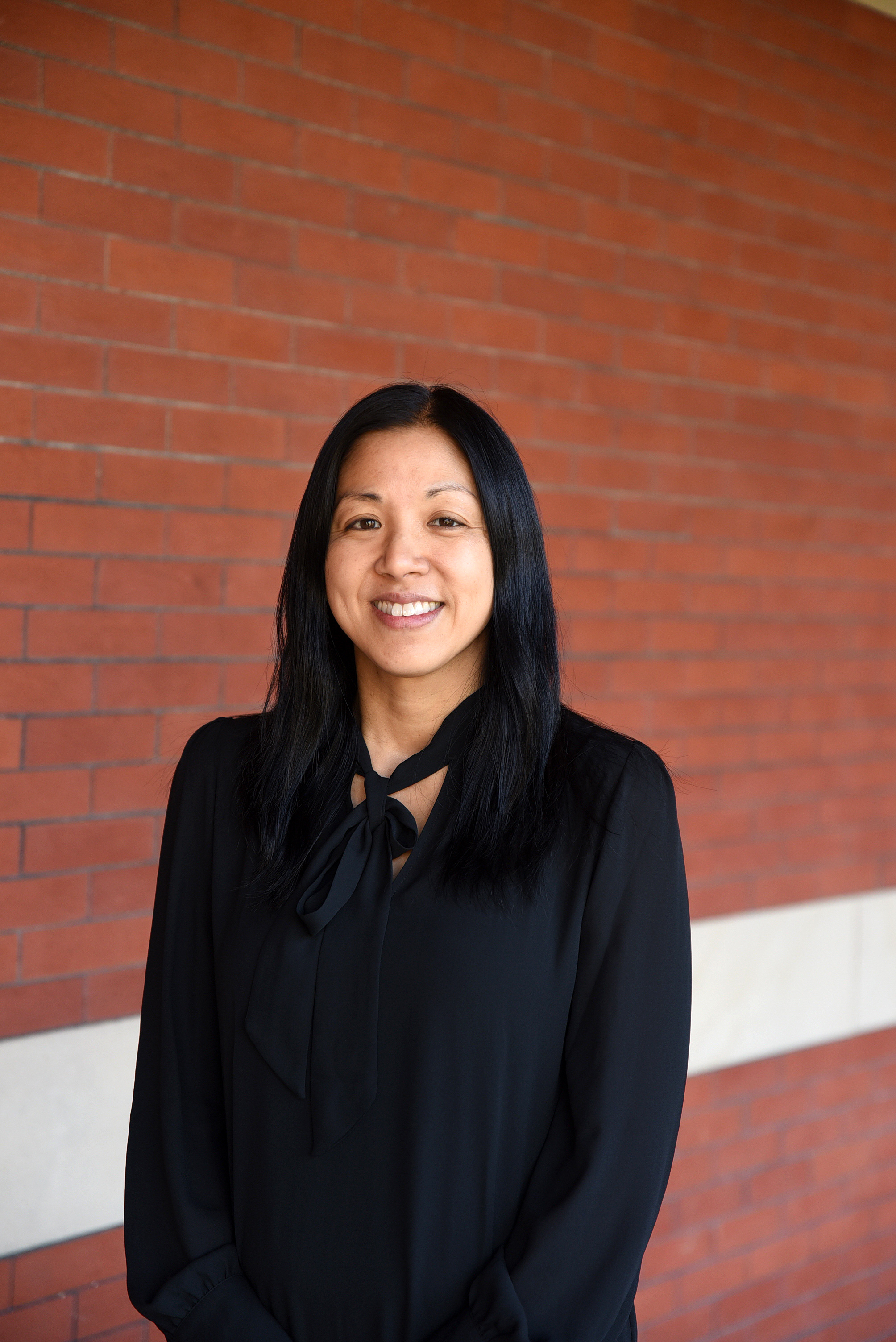 A picture of a smiling asian woman with a black suit standing in the front of a red brick wall