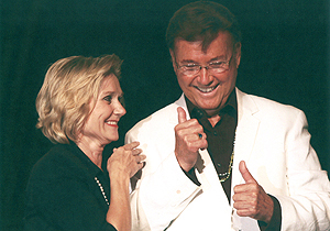 Bobbi Scoggins in a dark outfit stands next to Bernard Bragg, who's wearing a white suit jacket and is signing something.