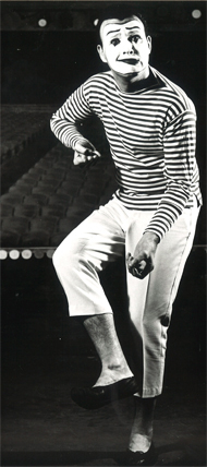 Bernard Bragg in striped shirt, white pants that mimes typically wear. His face has white makeup and made up eyebrows, eyes and lips, as mimes typically do. He's doing a mime pose, with a smirk like expression