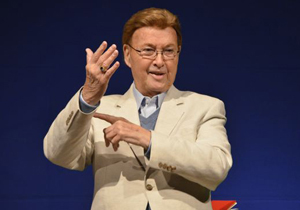 Bernard Brag stands to address an audience, with a beige-colored suit, gray v-neck sweater and shirt.