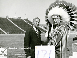 David Peikoff on a football field, in a suit and tie, standign next to a Native American Chief with a large headress and native garb. A square with the number 171 is below them and there is some printed text next to that. Text is not readily visible.