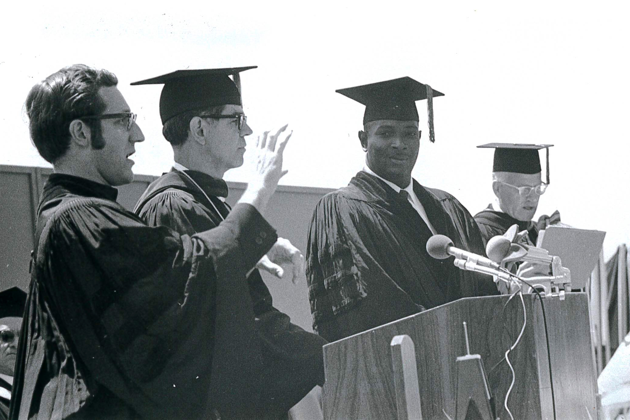 Andrew Foster in Doctoral regalia standing at the podium among three other white men including President Elstad.