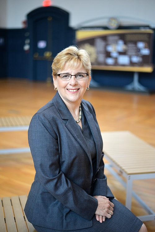 President Roberta J. Cordano, "Bobbi" sits on a bench with a blue-gray suit, in the Gallaudet Museum.