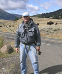 Robert E. Johnson, hoding a fishing rod off the road, in a dry landscape. Mountains are behind him.